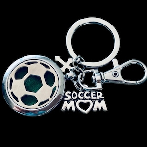 Puede incluir: Un llavero plateado con forma de balón de fútbol y un interior verde. El llavero incluye un llavero dividido, un mosquetón y un dije que dice "SOCCER MOM" con un símbolo de corazón.
