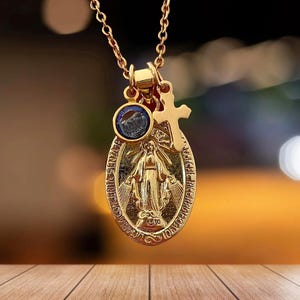Può includere: Collana con catena d'oro con un ciondolo ovale in oro che raffigura la Vergine Maria. Il ciondolo ha anche una piccola pietra preziosa blu e un ciondolo a forma di croce d'oro.