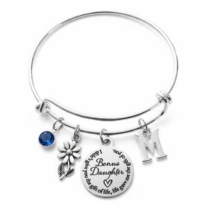 May include: Silver-toned bangle bracelet with multiple charms. Charms include a blue gemstone, a flower, a disc with the words "Bonus Daughter", and the letter "M". The bracelet is designed to be adjustable.