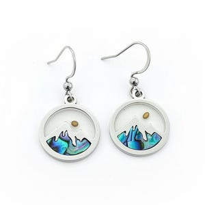 May include: A pair of silver-toned dangle earrings. Each earring features a circular pendant with a mountain scene. The mountains are made of iridescent abalone shell, and a small gold-colored sun is above them. The earrings hang from silver-colored hooks.