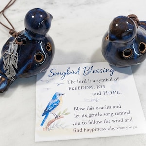 Könnte beinhalten: Zwei keramische, vogelförmige Okarinas in dunkelblauer Glasur, mit Federanhänger und brauner Kordel. Eine Karte mit der Aufschrift "Songbird Blessing" mit Text über Freiheit, Freude und Hoffnung. Die Okarinas haben drei Löcher.