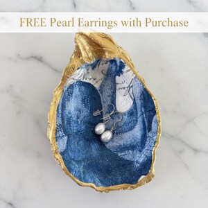 Op de afbeelding: Een decoratieve oesterschelp met een goudkleurige rand en een blauwe octopusprint aan de binnenkant. De schelp bevat een paar zilverkleurige oorbellen met witte parels. De tekst FREE Pearl Earrings with Purchase is zichtbaar.
