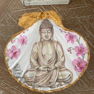 Puede incluir: Una concha de vieira decorativa con una ilustración pintada a mano de un Buda sentado en postura de meditación. La concha está decorada con flores de orquídea rosas y una pequeña pila de piedras grises, con los bordes pintados en oro metálico.