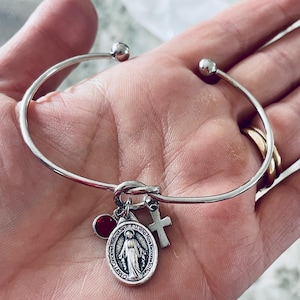 Może przedstawiać: Srebrna bransoletka typu bangle z węzłem i wiszącymi zawieszkami. Zawieszki obejmują czerwony kamień szlachetny, krzyż i owalny wisiorek z wizerunkami religijnymi i tekstem. Bransoletka ma prosty, elegancki design.