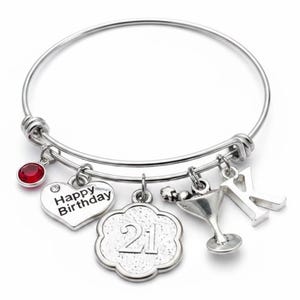 May include: A silver-toned expandable bangle bracelet featuring five charms: a round red crystal, a heart-shaped charm with the text Happy Birthday, a flower-shaped charm with the number 21, a martini glass charm, and a silver letter K charm.