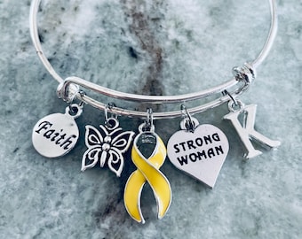 May include: A silver bangle bracelet with multiple charms. Charms include a round charm with the word "Faith", a butterfly, a yellow ribbon, a heart with "Strong Woman", and the letter "K". The bracelet is on a gray surface.