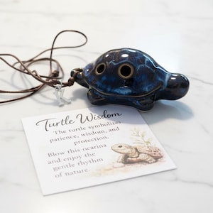 Könnte beinhalten: Blau-braune Keramik-Okarina in Schildkrötenform mit brauner Schnur. Daneben eine Karte mit dem Text "Turtle Wisdom" und einer Schildkröten-Illustration. Ein Musikinstrument.