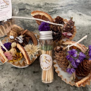 May include: A collection of fire starters with dried flowers, pinecones, and orange slices. A small glass vial of matches with a "Thank You" sticker is in the center. The starters are in decorative paper cups with wicks.