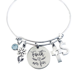 May include: Silver-toned bangle bracelet with multiple charms. Charms include a blue gemstone, a daisy, a cross with a heart cutout, and the letter "S". A round charm displays the phrase "Faith over fear" in script.