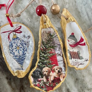 May include: Three Christmas ornaments made from oyster shells. Each shell has a gold painted edge and a different festive design. One features a blue and white ornament, another a winter scene with puppies, and the last a snowman.