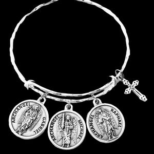 May include: Silver bangle bracelet with three round charms and a cross pendant. The charms depict archangels Gabriel, Michael, and Raphael. The bracelet has a hammered texture and a clasp closure, against a black background.