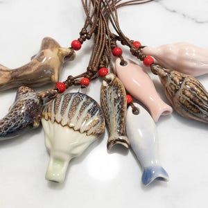 May include: A collection of ceramic whistles in various shapes, including fish and bird forms, strung on brown cords with red beads. The whistles are glazed in shades of brown, white, pink, and blue.