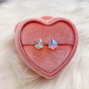 May include: A pair of iridescent, geometric stud earrings displayed inside a pink heart-shaped velvet jewelry box. The earrings have a multifaceted design, reflecting multiple colors. The box sits on a white, fluffy surface.