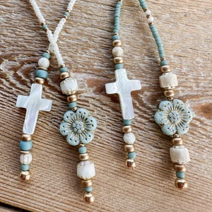 May include: Four beaded necklaces with a cross and flower pendant. The necklaces feature white, teal, and gold beads. The pendants are white with a cross shape and a light blue flower. The necklaces are on a wooden surface.
