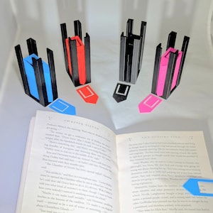 May include: Four 3D printed bookmark holders in black with different colored bookmarks. The bookmarks are blue, red, pink, and black. The holders are sitting on an open book.