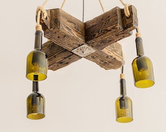 May include: A rustic chandelier crafted from reclaimed wood and repurposed wine bottles. The fixture features a cross-shaped wooden frame, rope suspension, and four green glass bottle lampshades with visible light bulbs. A decorative metal accent is centered on the wood.