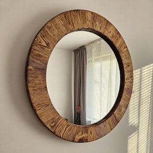 May include: A round mirror with a thick, rustic wooden frame. The frame has a natural wood finish with a dark brown stain. The mirror is reflecting a room with a window and blinds.