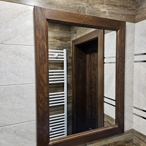 Puede incluir: Un espejo rectangular de baño con un marco de madera marrón oscuro. El espejo refleja un toallero blanco y una puerta marrón oscuro. Las paredes circundantes están alicatadas con una mezcla de baldosas claras y oscuras.
