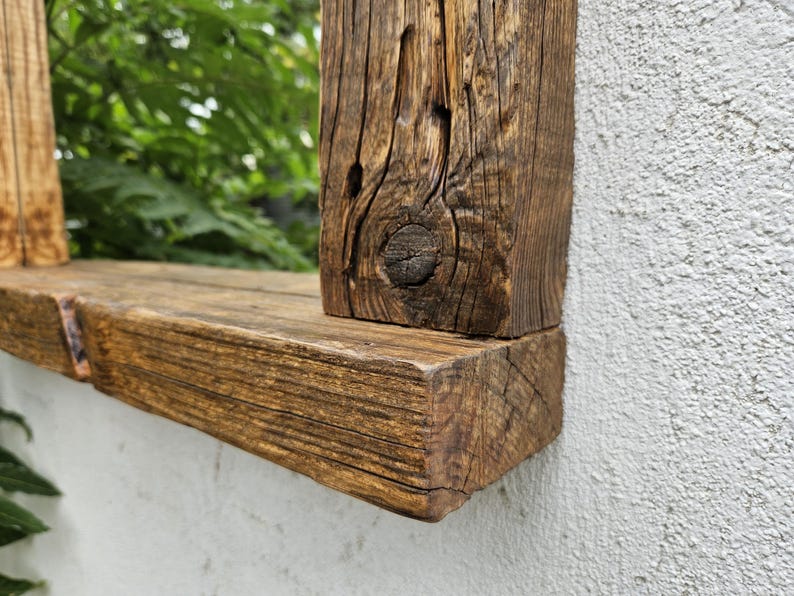 May include: Close-up of a rustic wooden mirror frame with a natural wood finish. The frame is made of thick, reclaimed wood planks with knots and grain patterns. The frame is mounted on a white wall.