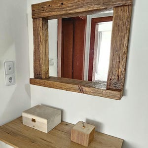 May include: A rustic wooden mirror with a shelf, featuring a natural wood finish and a rectangular shape. The mirror is mounted on a white wall above a wooden dresser with two drawers.