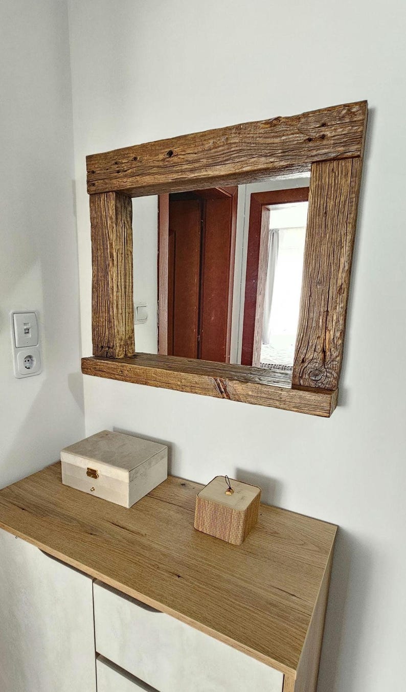May include: A rustic wooden mirror with a shelf, featuring a natural wood finish and a rectangular shape. The mirror is mounted on a white wall above a wooden dresser with two drawers.