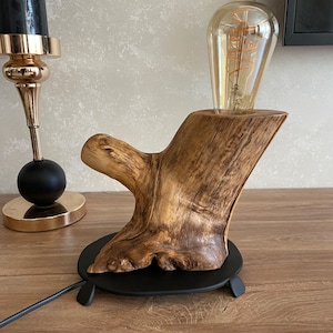 Puede incluir: Una lámpara de mesa de madera rústica con un acabado de madera natural. La lámpara presenta un diseño único en forma de rama y una base de metal negro. La lámpara está iluminada por una bombilla de vidrio transparente con un filamento en espiral.