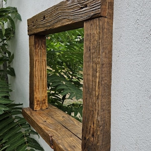 May include: A rustic wooden mirror with a shelf. The mirror is framed in reclaimed wood with a natural brown finish. The shelf is made of the same wood and is attached to the bottom of the frame.