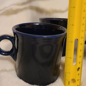 Może przedstawiać: Niebieski ceramiczny kubek z uchwytem, o wysokości około 9 cm. Kubek stoi na białym, puszystym podłożu obok linijki.