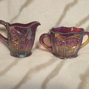 Puede incluir: Un juego vintage de jarra de crema y azucarero de vidrio rojo iridiscente con un patrón detallado. El juego está hecho de vidrio de carnaval.
