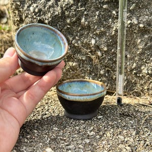 May include: Two small ceramic bowls with a dark brown matte exterior and a glossy, light blue interior glaze that transitions to a brown rim. A clear plastic pen is positioned vertically next to the bowls for scale, measuring 5.5 inches in length.