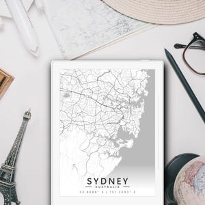 May include: A grayscale map of Sydney, Australia, displayed on a tablet. The image includes a miniature Eiffel Tower, a globe, a toy airplane, a pencil, eyeglasses, and a wooden box with letter stamps. The map has the city name and coordinates.