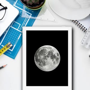 Può includere: Un tablet bianco con uno schermo nero che mostra un'immagine ravvicinata della luna. Il tablet è su una superficie bianca con vari articoli da ufficio, tra cui una calcolatrice, un righello, un metro a nastro, una matita e un tappetino blu a griglia.