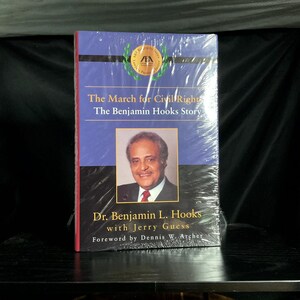 May include: A book titled "The March for Civil Rights: The Benjamin Hooks Story" is wrapped in clear plastic. The cover features a portrait of Dr. Benjamin L. Hooks, with text indicating the author and foreword writer. The book has a blue and red cover.