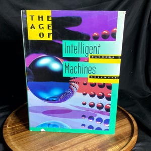May include: A hardcover book titled "The Age of Intelligent Machines" by Raymond Kurzweil. The cover features a vibrant design with the title in green and the author's name in yellow. The background includes spheres in blue, purple, and red.