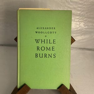 May include: A hardcover book with a bright green cover. The title "While Rome Burns" is printed in black, along with the author's name, Alexander Woollcott. The book is propped up on a wooden stand.