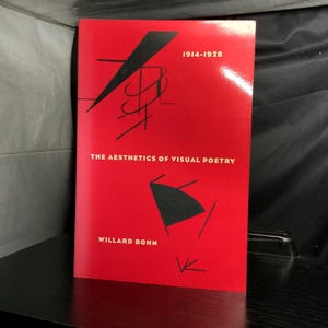 May include: A book titled "The Aesthetics of Visual Poetry" by Willard Bohn. The cover is red with black abstract shapes and the text "1914-1928". The book is standing upright on a black surface.