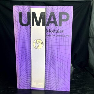 May include: A rectangular book titled UMAP Modules Tools for Teaching 1987 with a purple cover featuring a black grid pattern. A central white vertical strip displays a circular yellow and black globe illustration and the COMAP logo at the bottom.