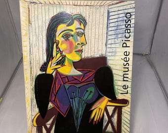 May include: A colorful painting of a seated woman in the style of Picasso, with the text "Le musée Picasso" on the side. The artwork features bold lines and vibrant colors, mounted on a wooden stand.