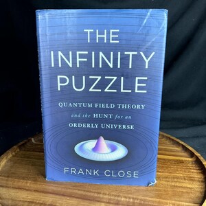 May include: A hardcover book titled The Infinity Puzzle: Quantum Field Theory and the Hunt for an Orderly Universe by Frank Close. The cover is blue with thin circular lines and a central graphic of a purple and white wave-like structure.