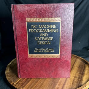 May include: A hardcover book titled NC Machine Programming and Software Design by Chao-Hwa Chang and Michel A. Melkanoff. The book features a textured, deep red cover with a central black rectangular panel containing gold-colored text.