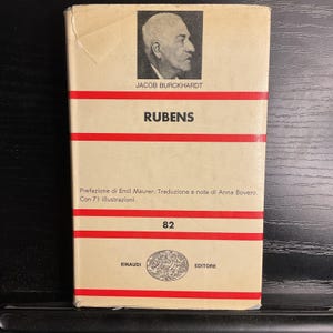 May include: A vintage book titled "RUBENS" by Jacob Burckhardt. The cover is beige with red horizontal stripes and features a portrait and text in Italian. The book is from the Einaudi Editore publishing house.