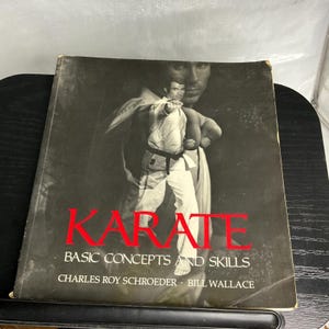 May include: A black and white instructional book titled KARATE BASIC CONCEPTS AND SKILLS by Charles Roy Schroeder and Bill Wallace. The cover features a large, faded portrait of a person in a karate uniform with a black belt in a fighting stance.