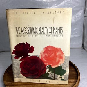 May include: A hardcover book titled The Algorithmic Beauty of Plants by Przemyslaw Prusinkiewicz and Aristid Lindenmayer. The cream-colored cover features two dark red roses and one light pink rose, with the text Springer-Verlag at the bottom.