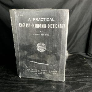 May include: A black hardcover book titled A Practical English-Mandarin Dictionary by Chiang Ker Chiu. The cover features white text, a circular geometric emblem, and the Chin Fen Book Store address in Singapore printed at the bottom.