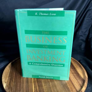 May include: A hardcover book titled "The Business of Investment Banking" in a teal cover with gold lettering. The book is a second edition and includes the author's name, K. Thomas Liaw, and the subtitle "A Comprehensive Overview."