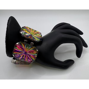 May include: A silver-toned bracelet with two large, rectangular iridescent crystals. The crystals display a vibrant array of colors, including pink, yellow, green, and blue. The bracelet is displayed on a black hand-shaped form.