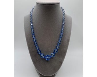 May include: A blue beaded necklace displayed on a gray mannequin. The necklace features faceted, round beads in varying sizes, with a larger, central focal bead. The beads are strung together, creating a sparkling, elegant accessory.