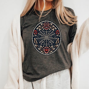 May include: Dark grey long-sleeved top with a circular design featuring a spiderweb, roses, and leaves. The design is in red, gold, and cream. The shirt is made of a soft material.