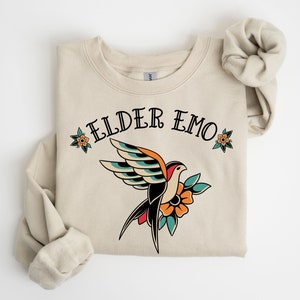Op de afbeelding: Een beige sweatshirt met de tekst "ELDER EMO" in zwart gedrukt. Een gestileerde tattoo-achtige vogel met kleurrijke vleugels en een bloem is onder de tekst gedrukt.
