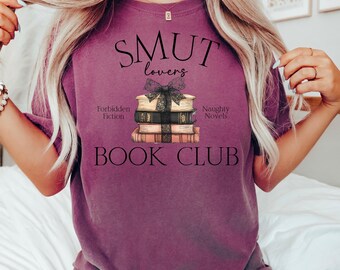 Op de afbeelding: Een paars T-shirt met de tekst "SMUT lovers BOOK CLUB" en een stapel boeken met een zwart lint en strik. De boeken hebben de tekst "Forbidden Fiction" en "Naughty Novels" erop.